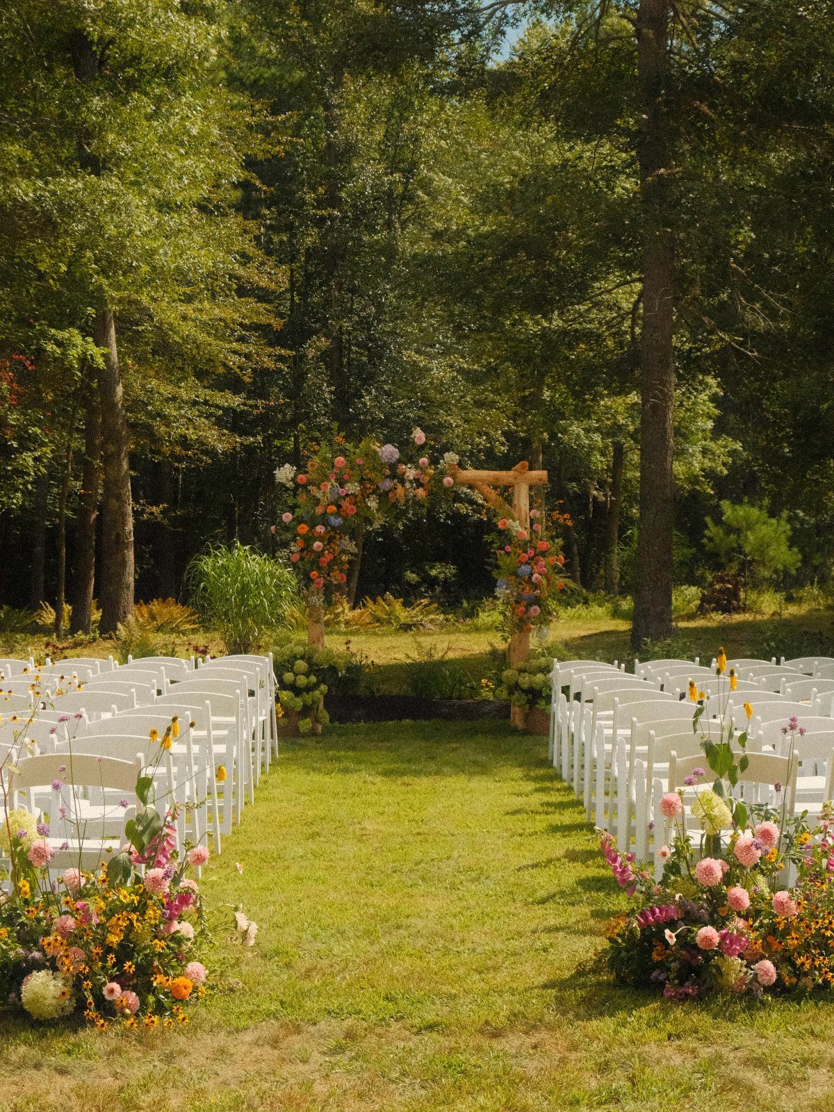 White wedding chairs arranged in rows on a lawn leading to a floral wooden ceremony arch.