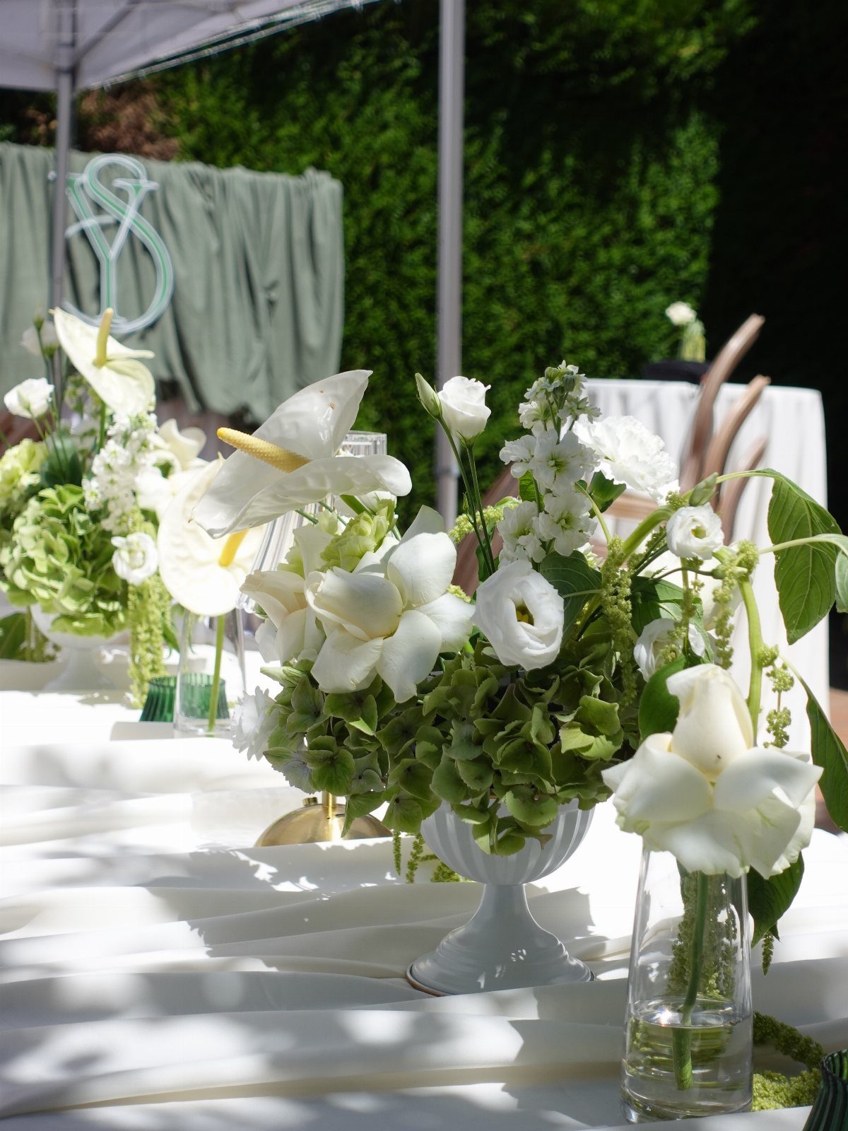 White floral arrangements in various vases arranged on a white tablecloth in a garden setting.