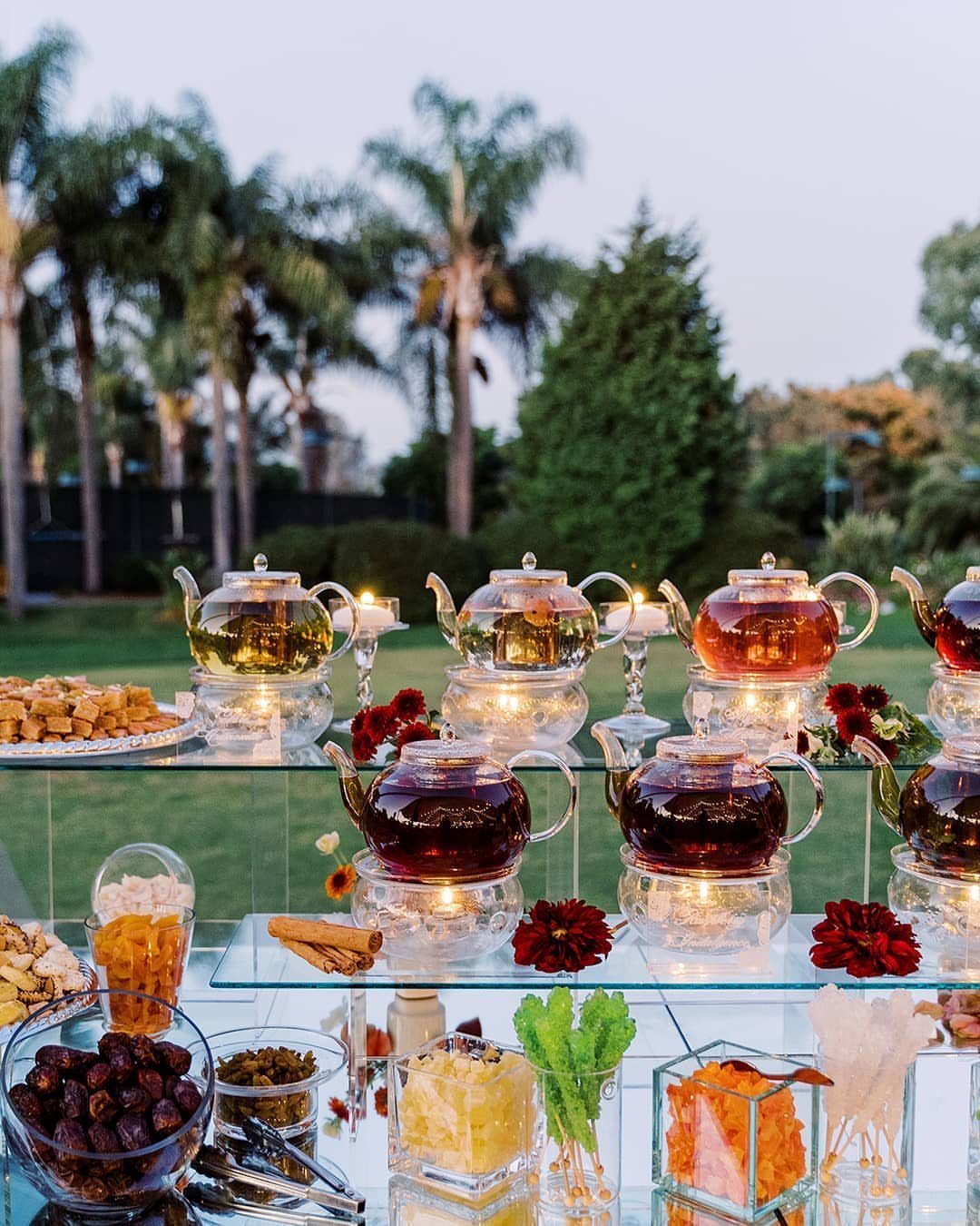 Glass teapots filled with colorful liquids sit on a tiered glass table outdoors.
