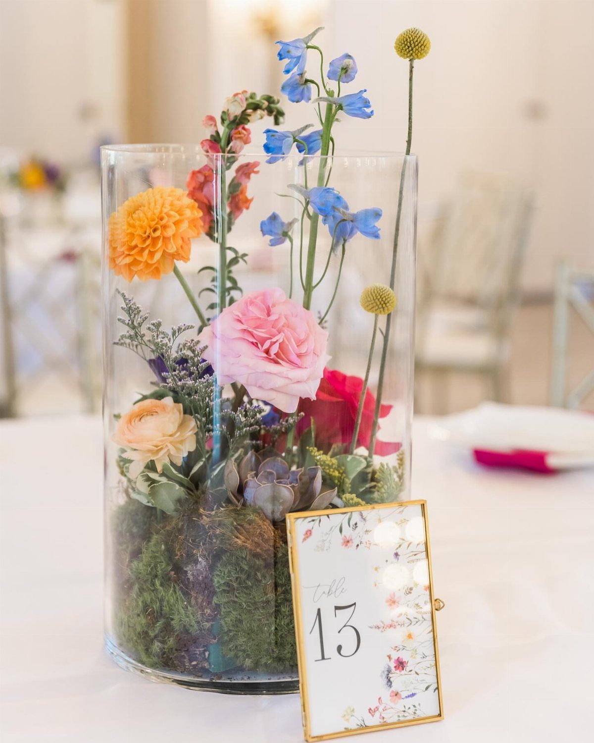 A glass vase filled with colorful garden flowers and green moss sits on a white table.