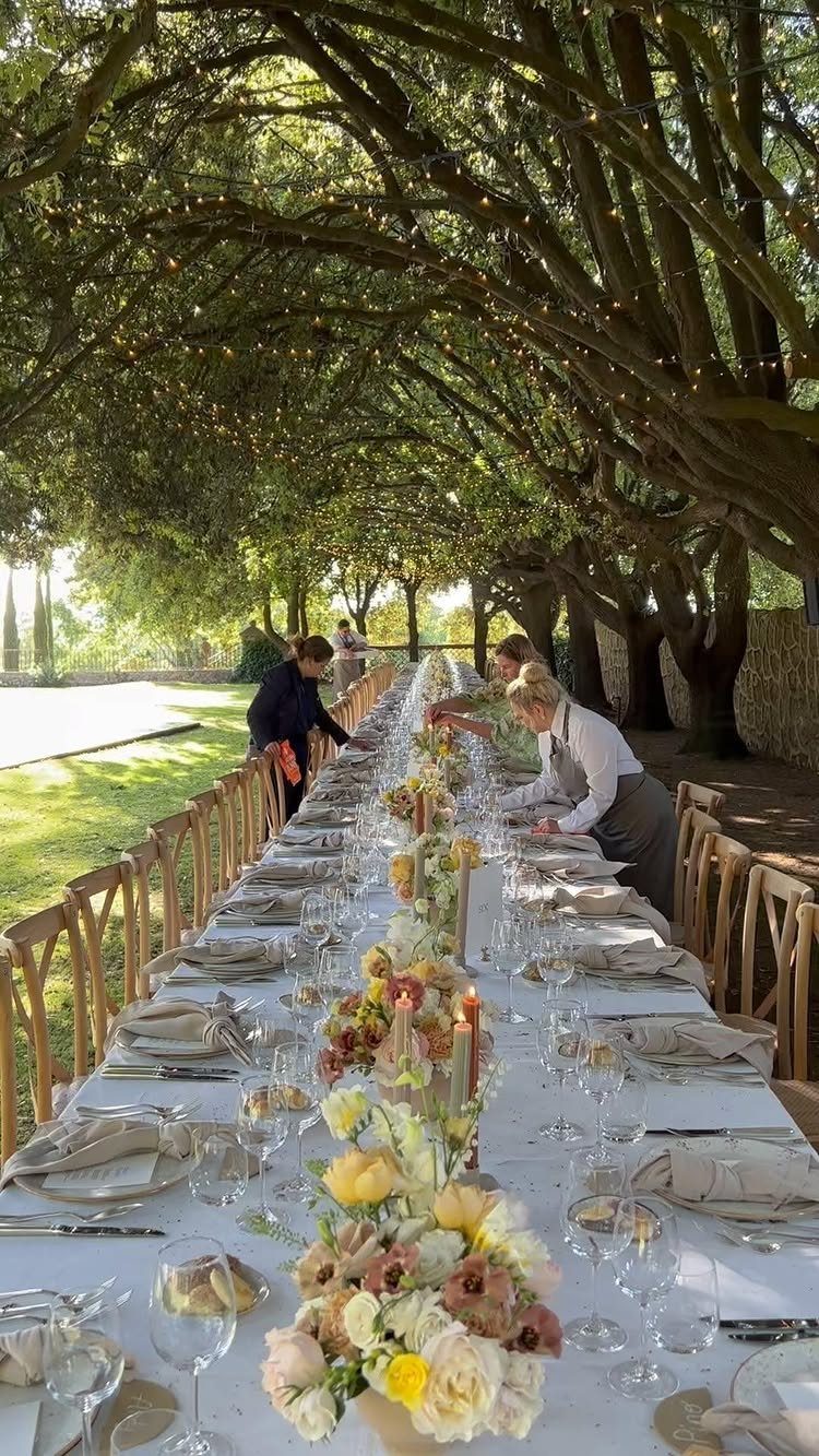 A long dining table set with flowers and glassware under a canopy of trees.