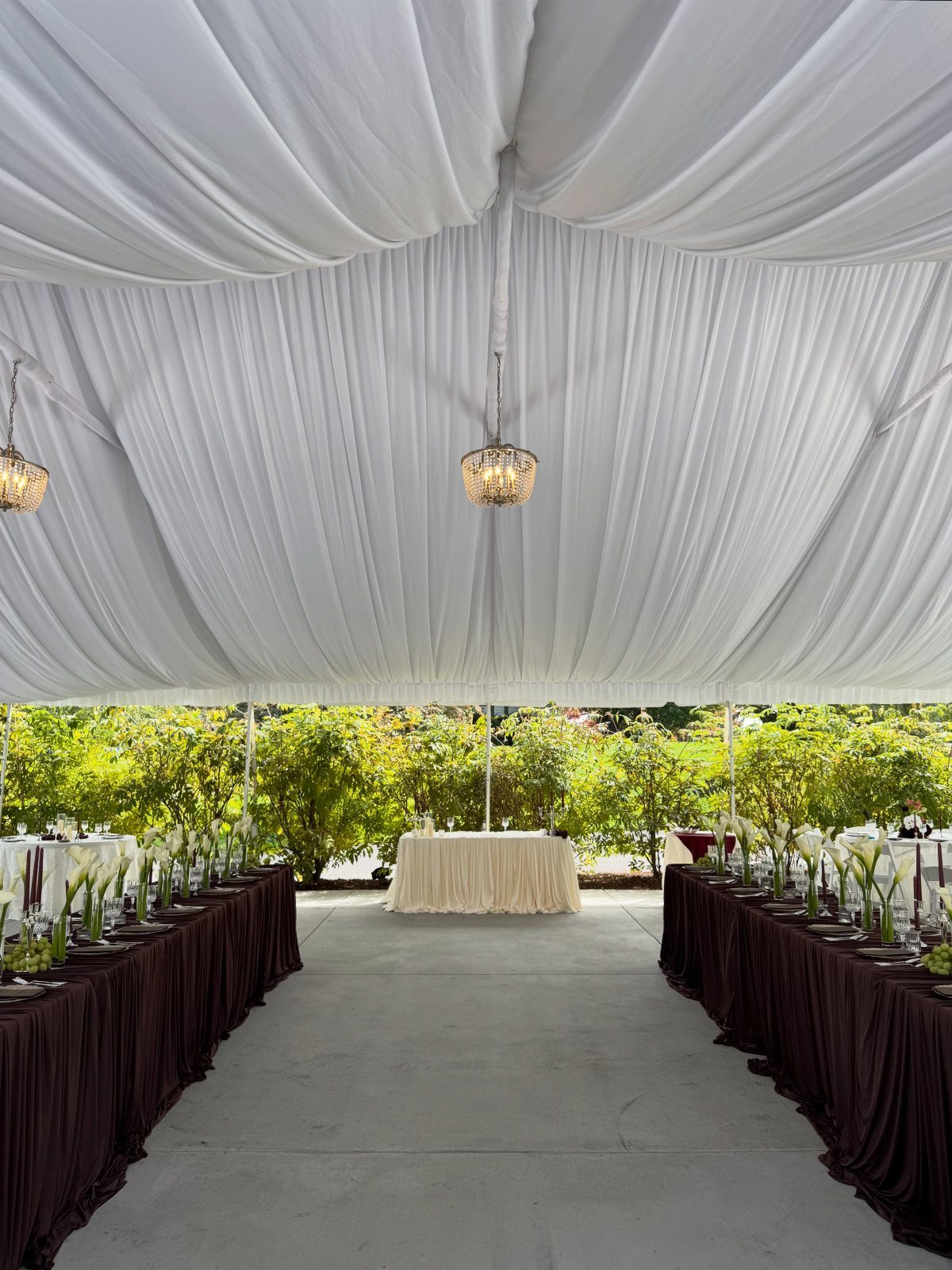 White draped wedding tent interior with long banquet tables and floral centerpieces overlooking a green garden.