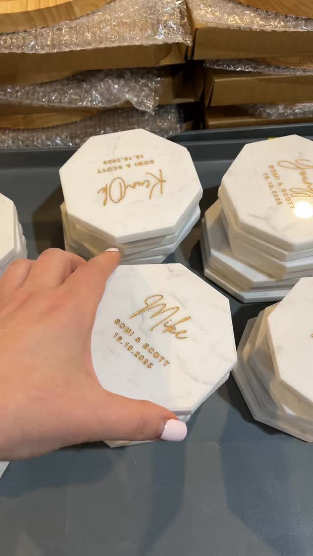 A hand holds a white marble hexagonal coaster with gold lettering and names.
