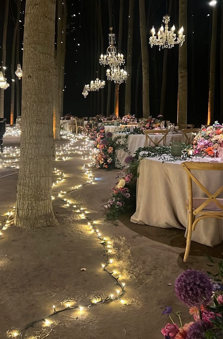 Wedding tables with floral arrangements and chandeliers under trees with string lights on the ground.