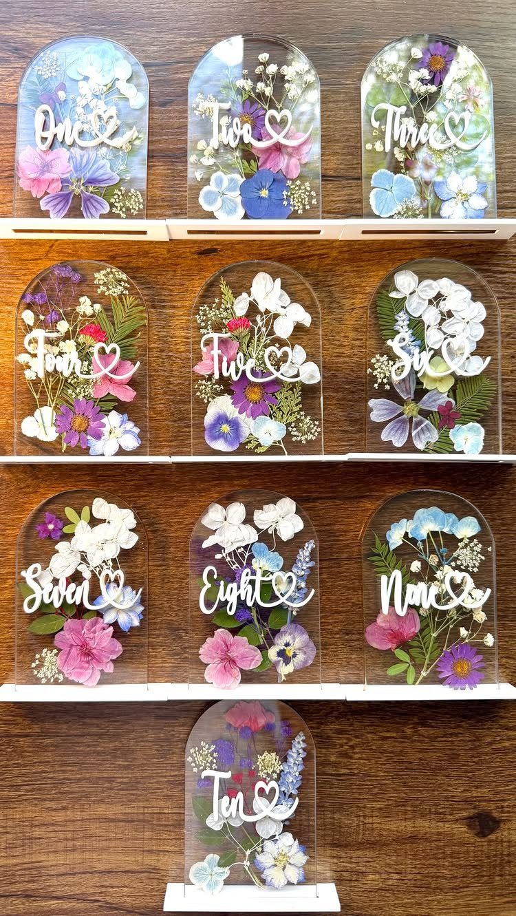 Ten arched acrylic table numbers containing colorful pressed flowers arranged on a wooden surface.
