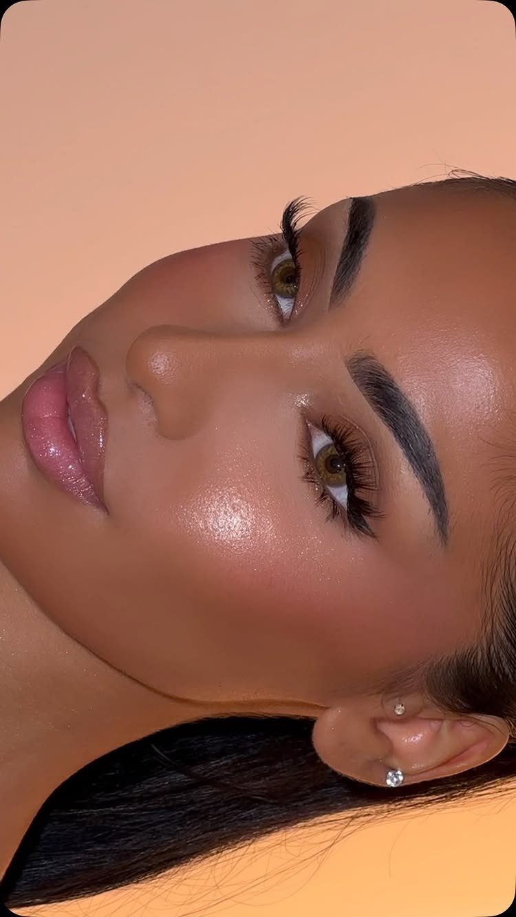 A woman with glowing skin, long lashes, and shiny pink glossy lips against a peach background.