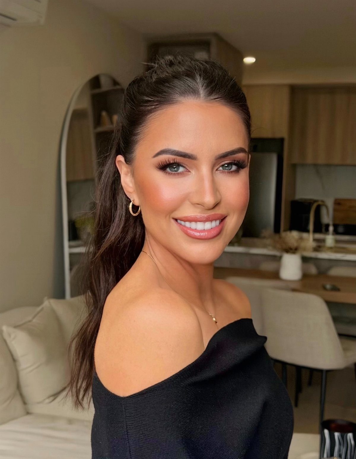 A smiling woman with long dark hair and radiant, glowing makeup looking towards the camera.