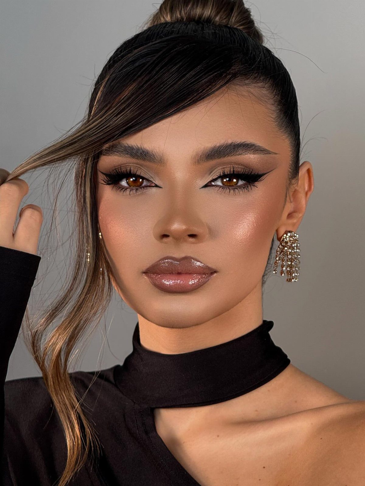 Woman with dramatic winged eyeliner, shimmery eyeshadow, and sculpted brows posing against a grey background.