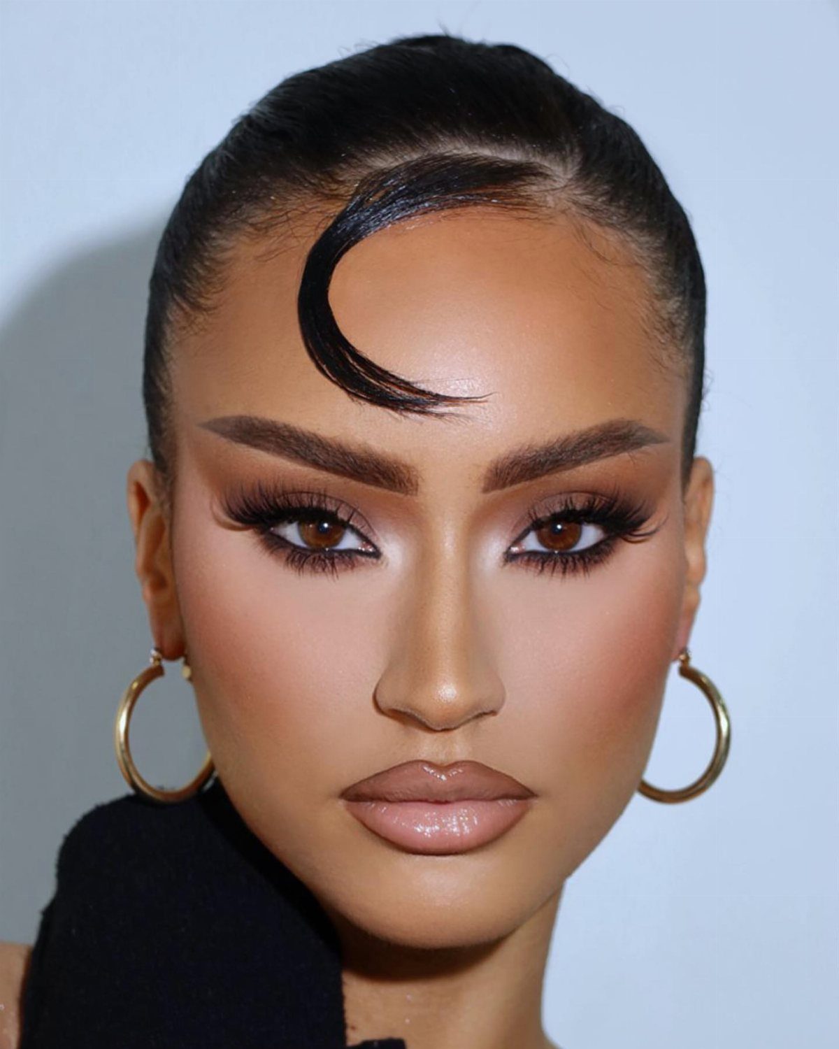 Woman with heavy glam makeup, voluminous lashes, and sculpted cheekbones wearing gold hoop earrings.