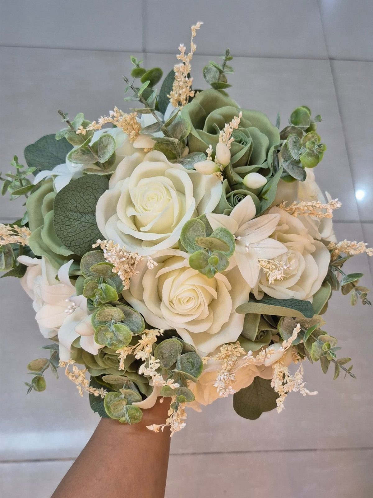 A bridal bouquet with ivory roses, white lilies, and green succulents held against a tiled floor.