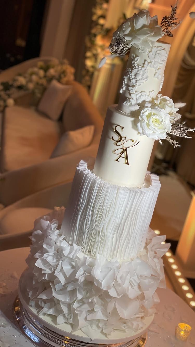 An ivory tiered wedding cake decorated with white ruffles, floral frosting, and gold monogrammed initials.