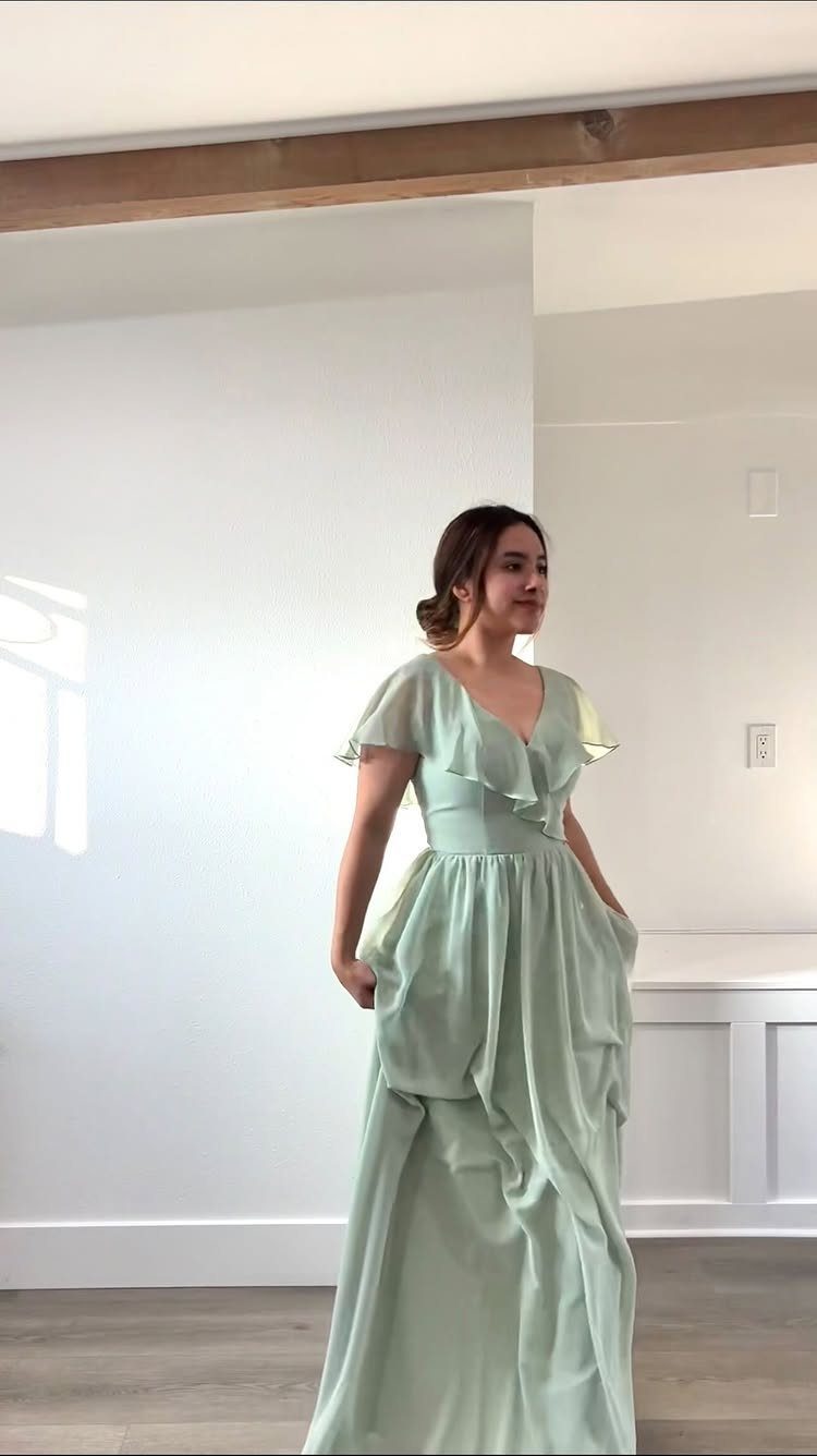 A woman posing in a long, flowing sage green bridesmaid dress against a white wall.