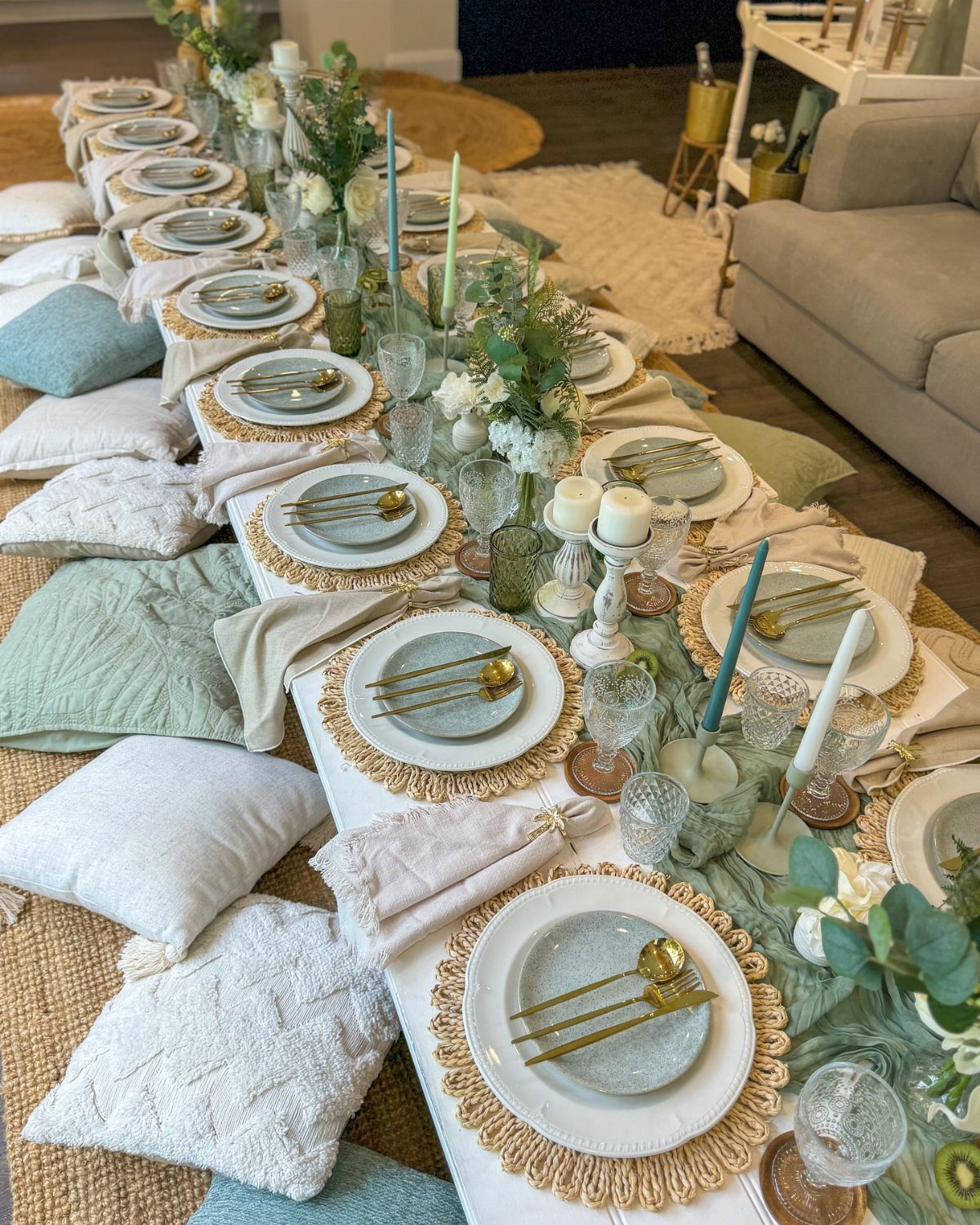 A long dining table decorated with sage green plates, gold cutlery, greenery, and ivory accents.