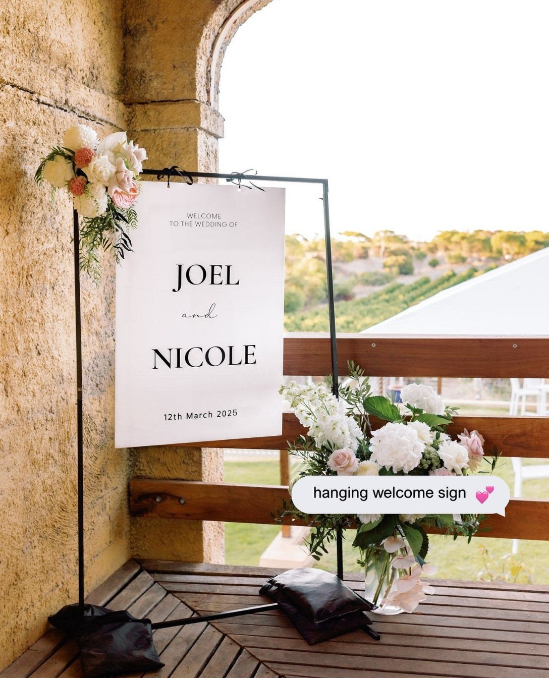 A white wedding welcome sign with floral arrangements on a black metal stand outdoors.