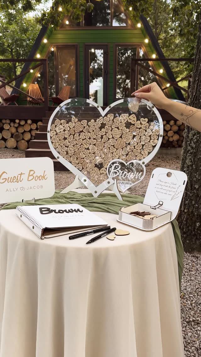 A hand placing a wooden heart into a large clear heart-shaped guest sign-in frame.