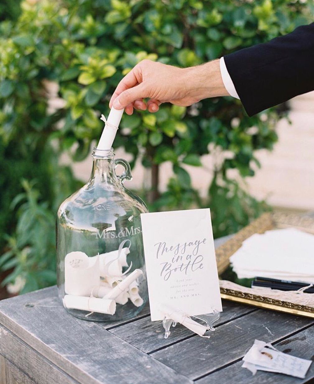 A hand placing a rolled paper message into a large glass bottle labeled Mrs. and Mrs.