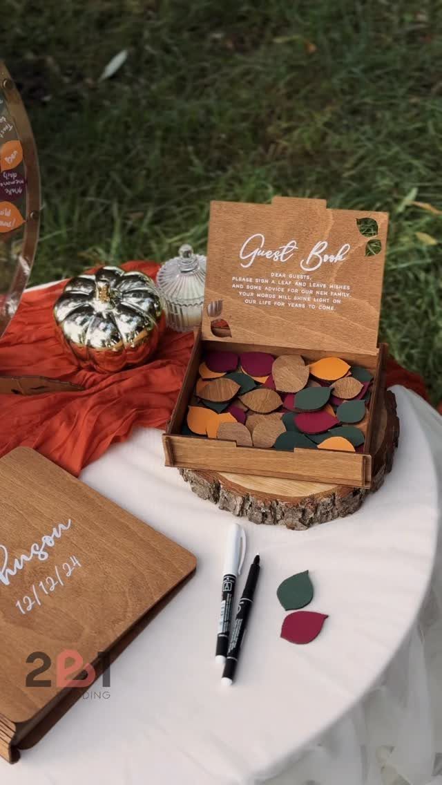 Wooden guest book box filled with colorful leaf-shaped cards on a white table outdoors.