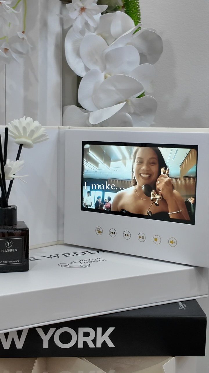 A white digital photo frame displaying a video of a smiling woman on a white table.