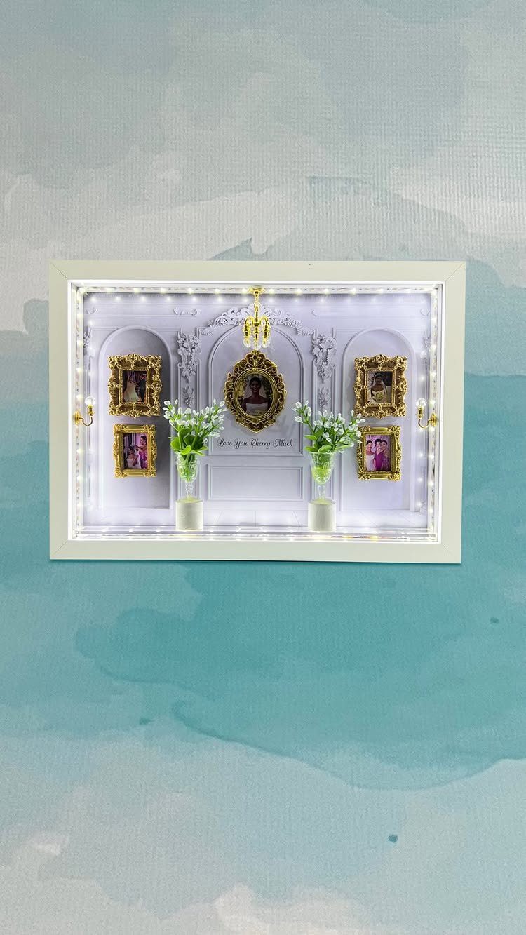 A white shadow box frame decorated with small gold photo frames and tiny white lights.