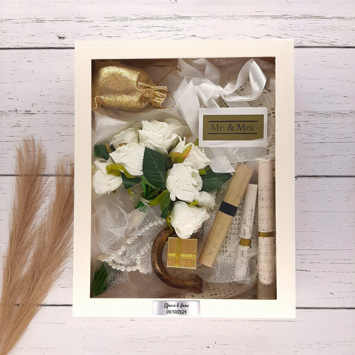 A white shadow box filled with white roses, scrolls, and wedding keepsakes on a wooden surface.