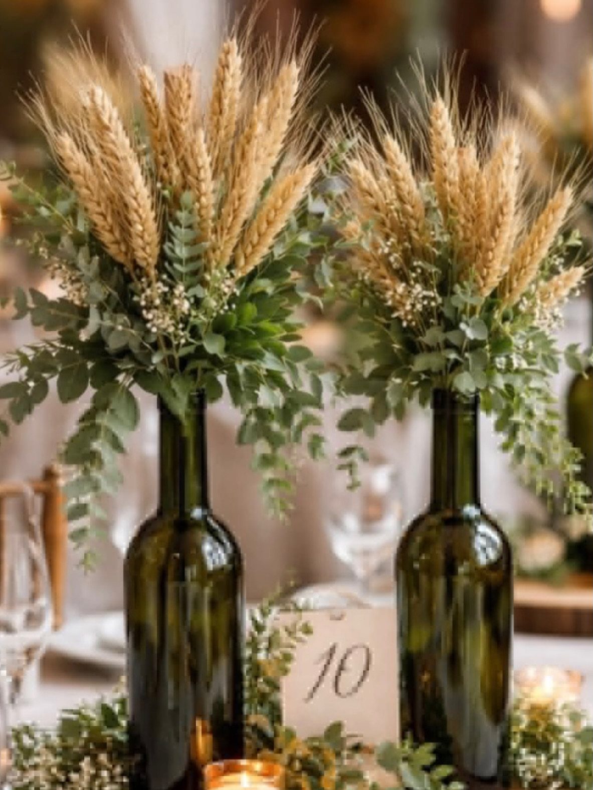 Two green glass bottles filled with wheat stalks, eucalyptus greenery, and small white flowers on a table.