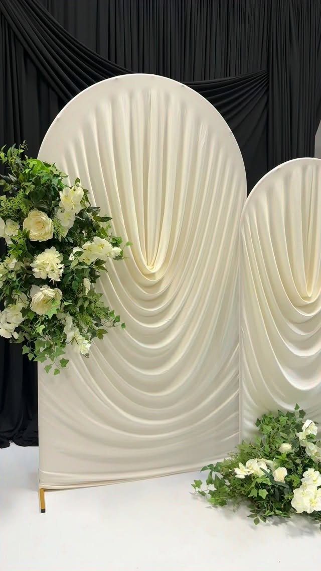 Cream pleated fabric arched backdrops decorated with white roses and green leaves against black curtains.