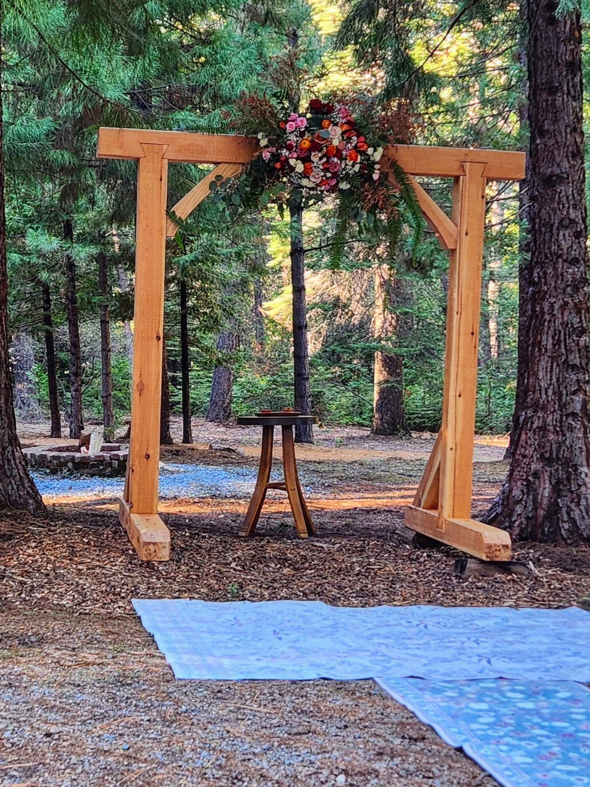 A wooden wedding arbor decorated with colorful flowers stands in a sunlit forest clearing.