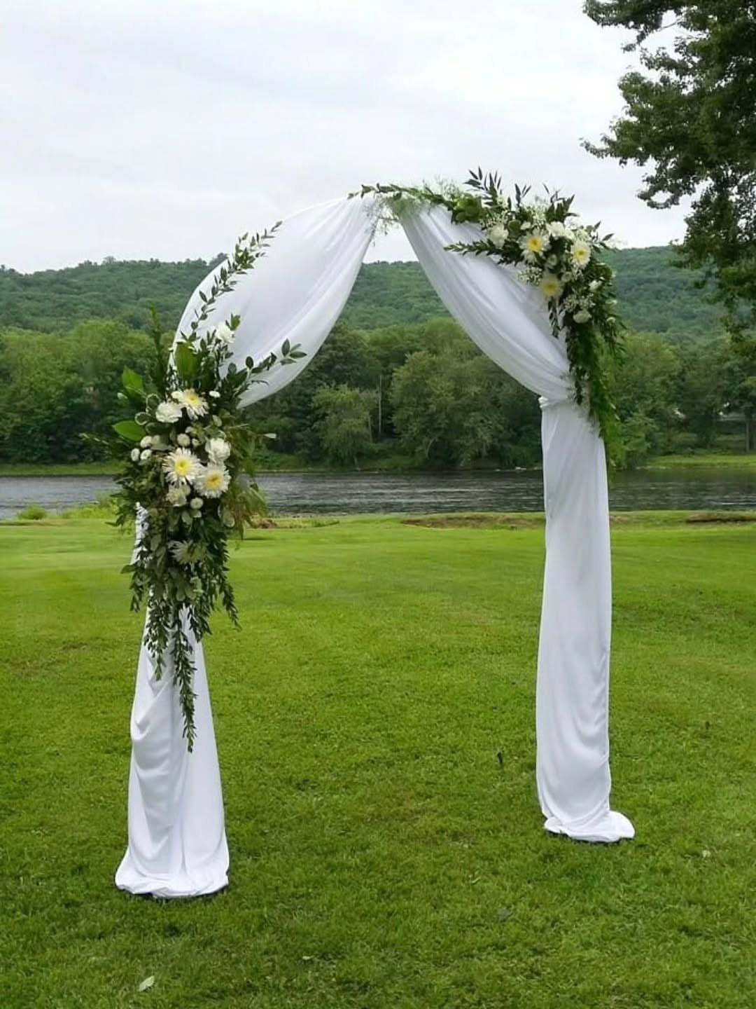 White fabric wedding arch decorated with green leaves and white flowers on a grassy field.