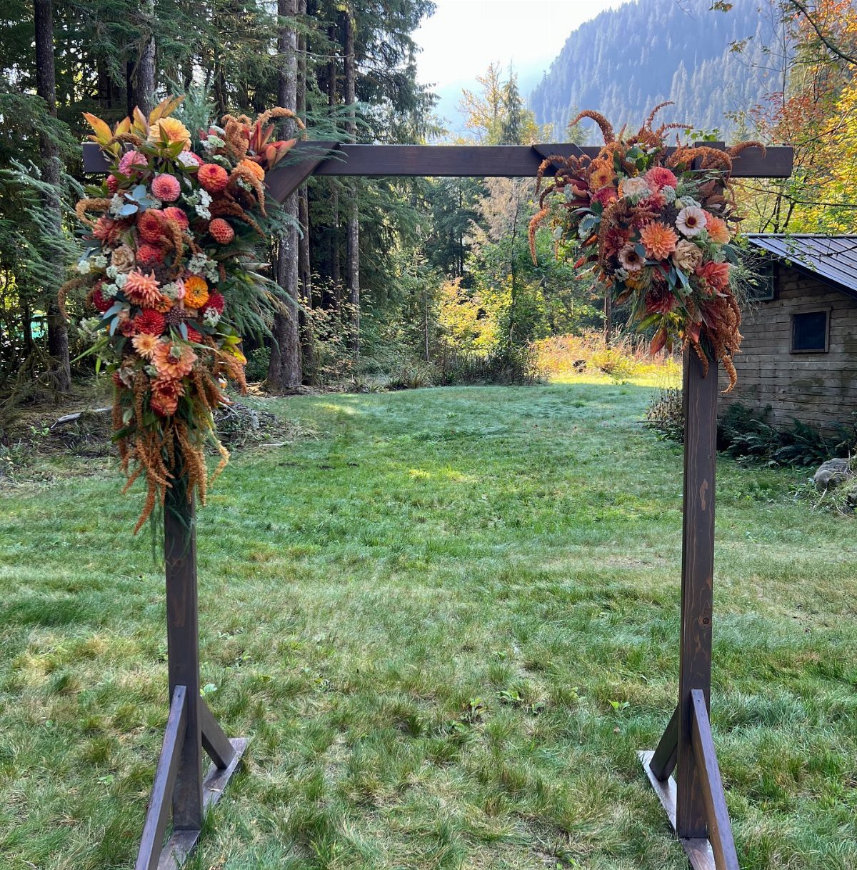 A dark wooden wedding arbor decorated with autumn floral arrangements in a grassy forest clearing.