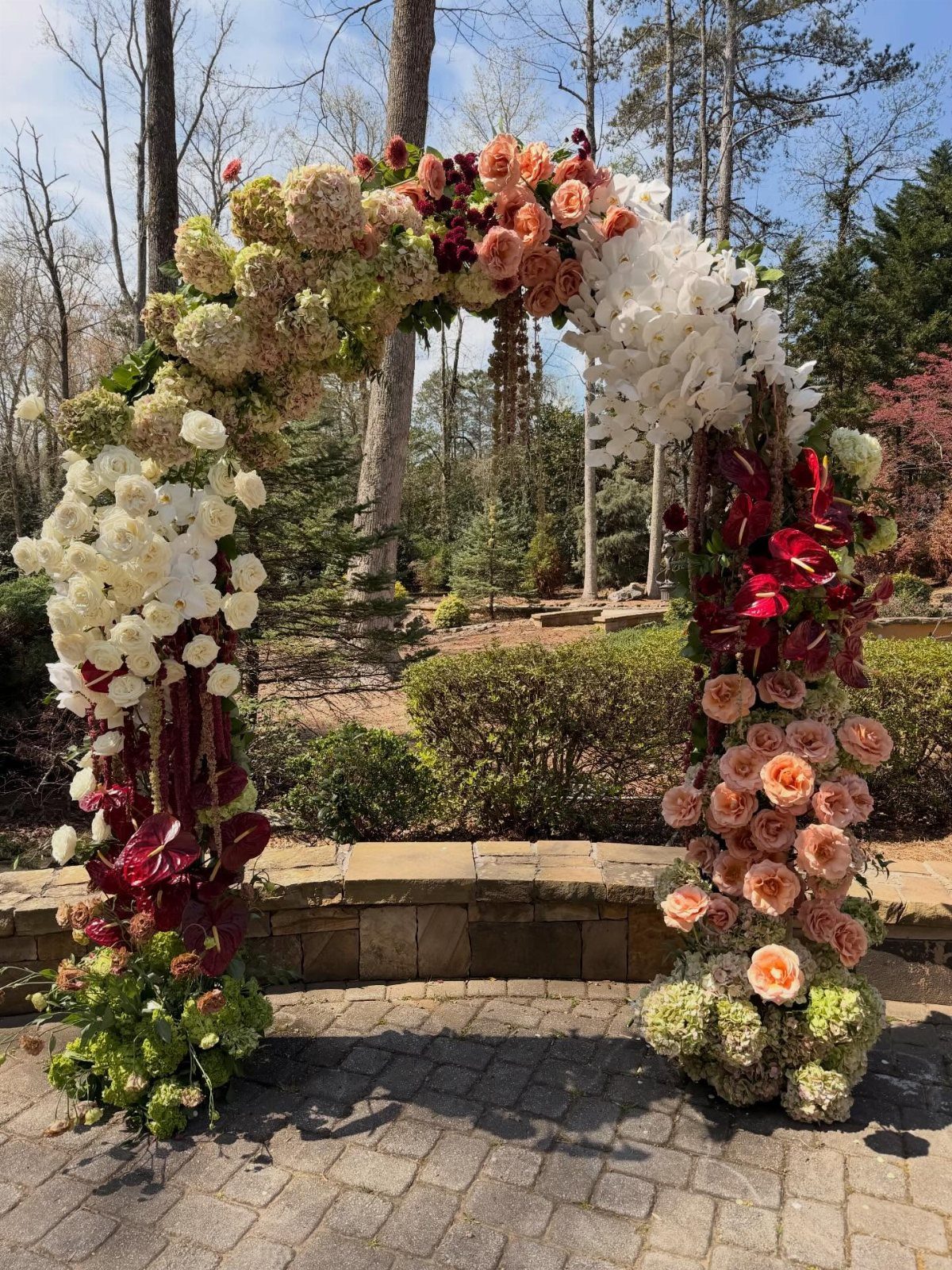 A lush floral wedding arch featuring white, peach, and red flowers set against a wooded background.