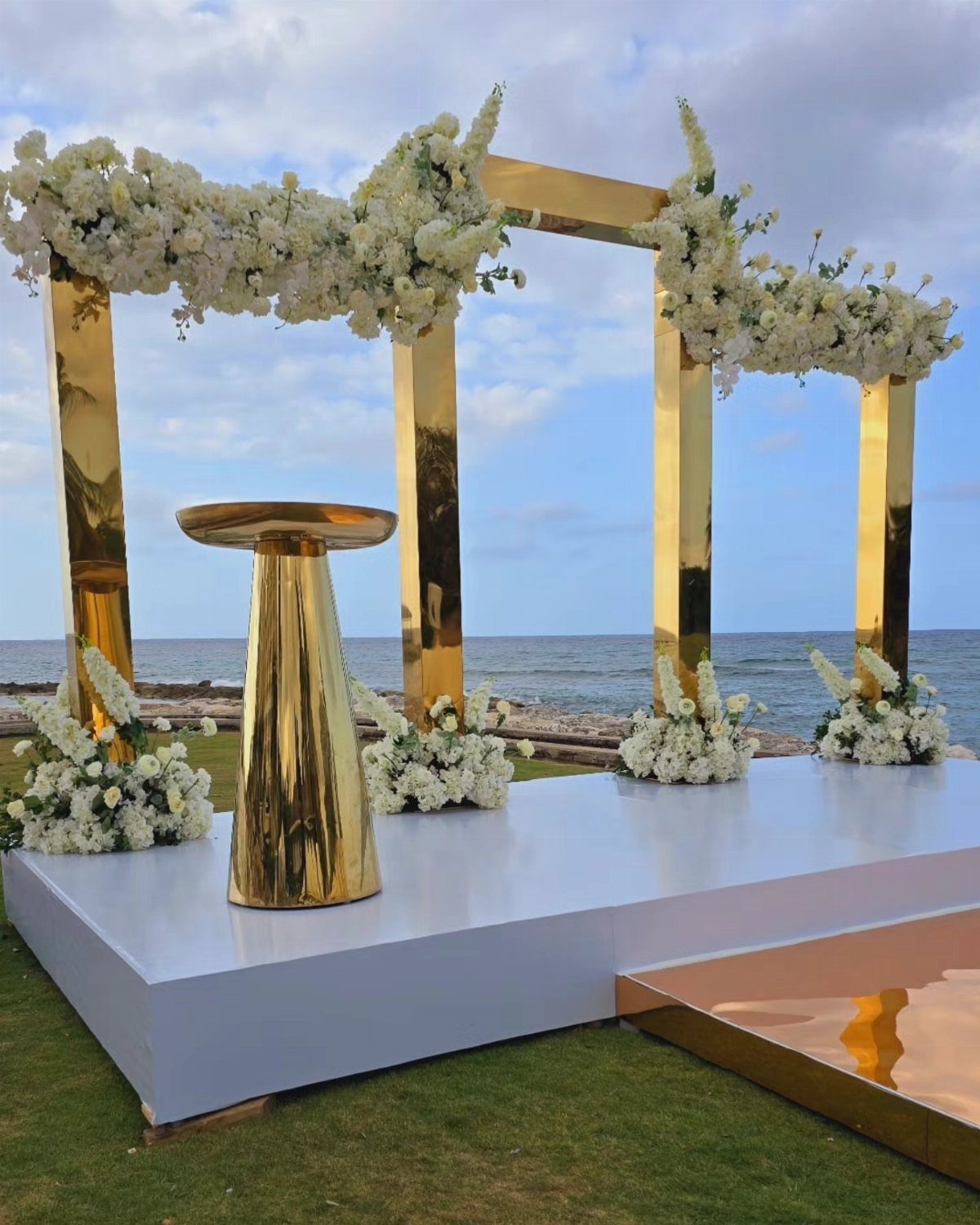 Gold geometric wedding arch decorated with white flowers on a platform by the sea.