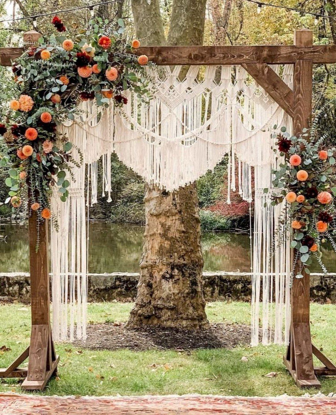 Wooden wedding arch with white macrame hanging and orange floral arrangements in a garden setting.