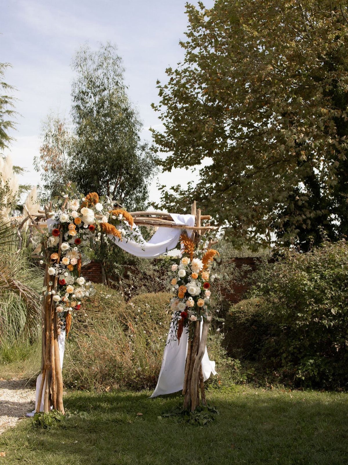 A natural wood branch wedding arch decorated with white fabric and orange flowers in a garden.
