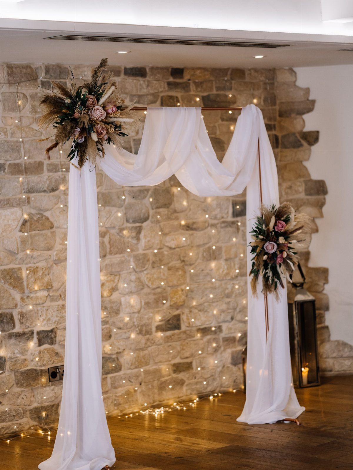 A copper pipe wedding arch draped in white fabric with dried flowers against a stone wall.