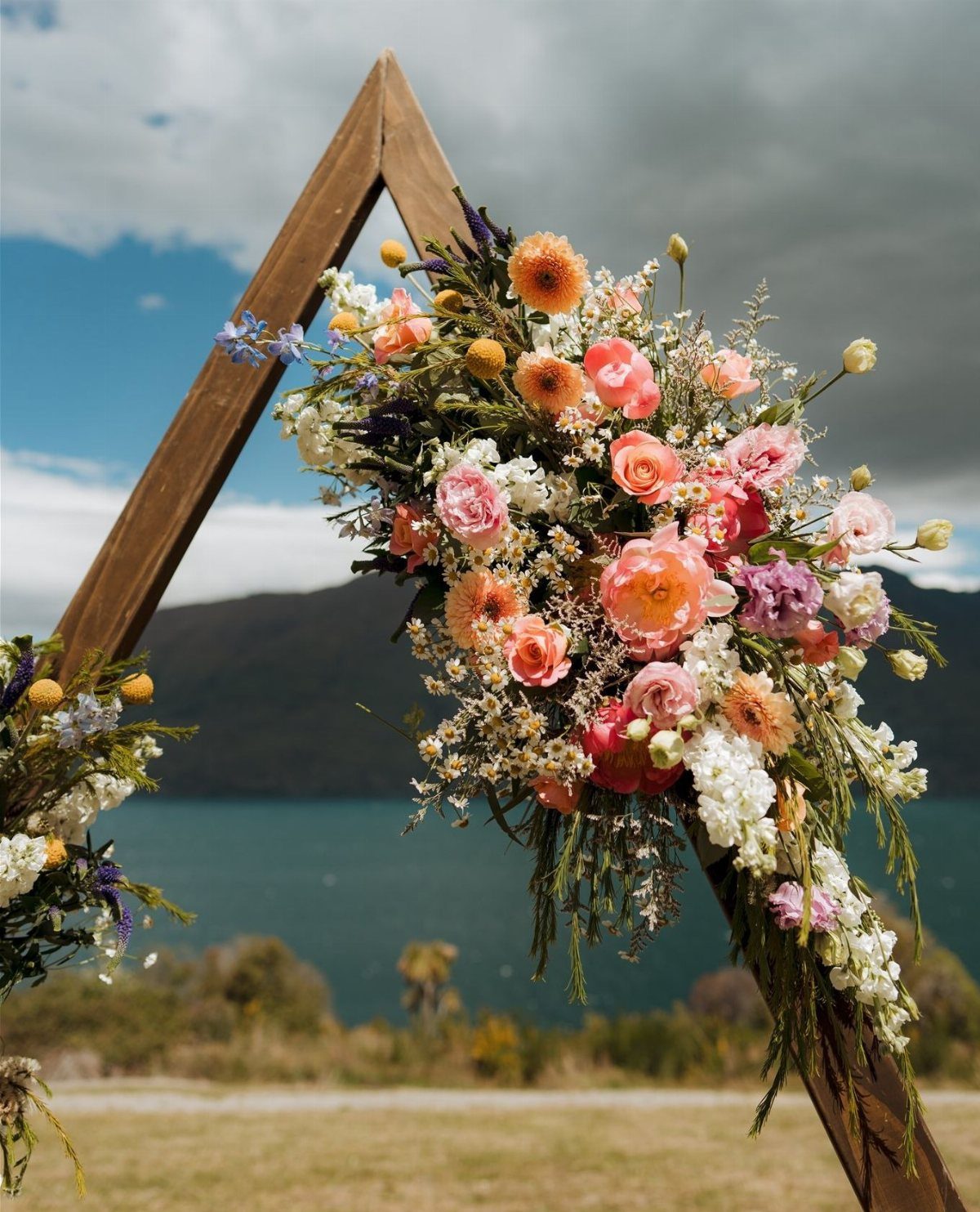 A wooden triangular wedding arch adorned with colorful peach and pink floral arrangements overlooking a lake.