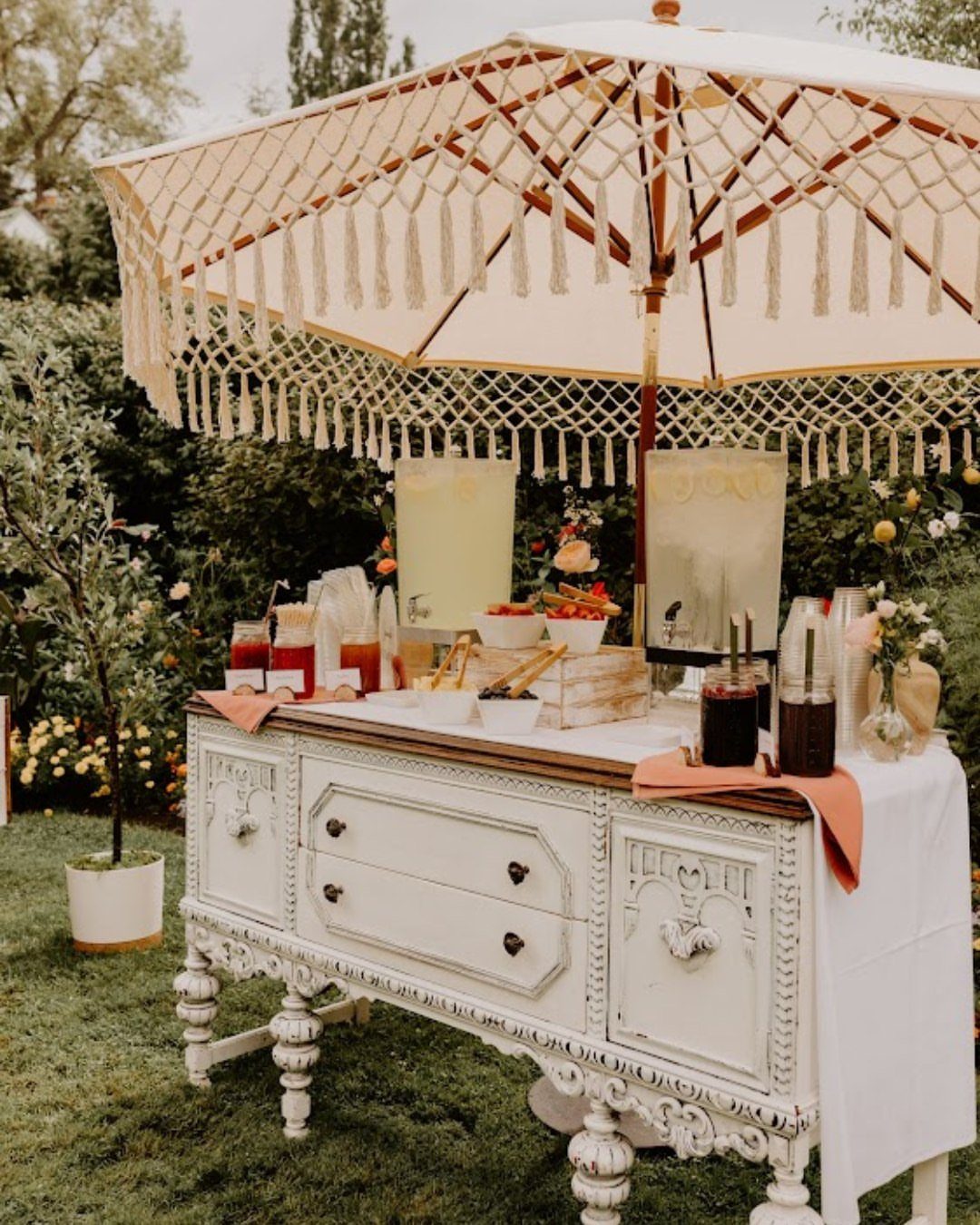A white vintage sideboard serving as an outdoor beverage station under a macrame umbrella.