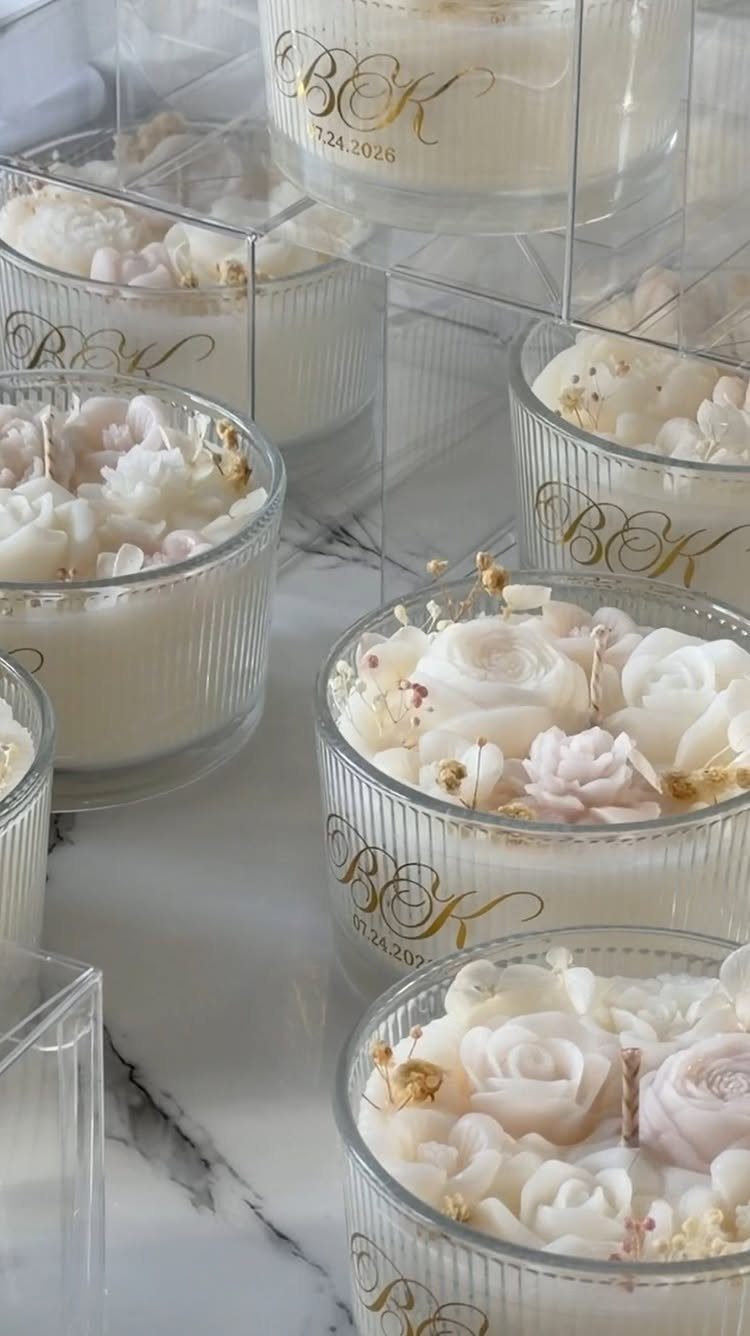 Several white rose-shaped wax candles in clear glass jars with gold monogramming on marble.