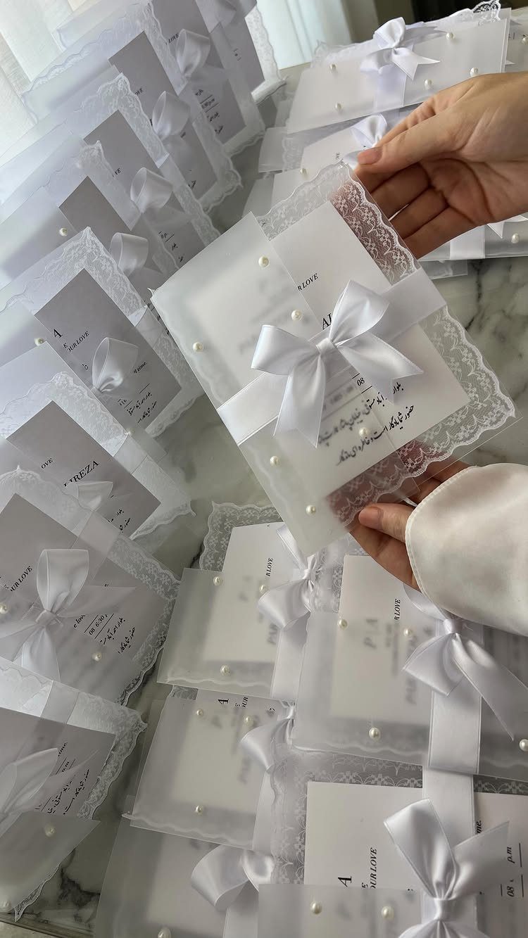 Hands holding a white lace wedding invitation decorated with a satin bow and pearls.