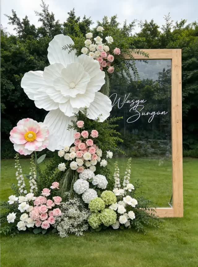 Large white paper flowers and pink floral arrangements decorating a wooden frame backdrop on a lawn.