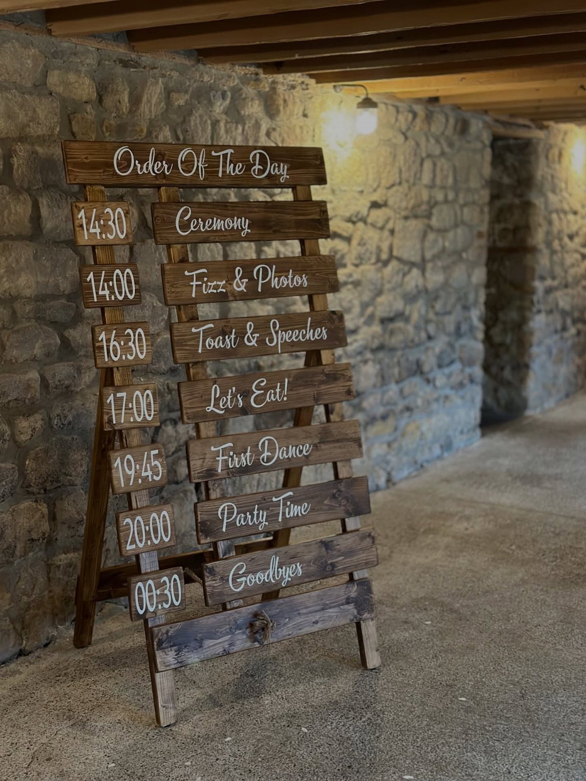 A rustic wooden plank sign displaying a wedding order of the day schedule against a stone wall.