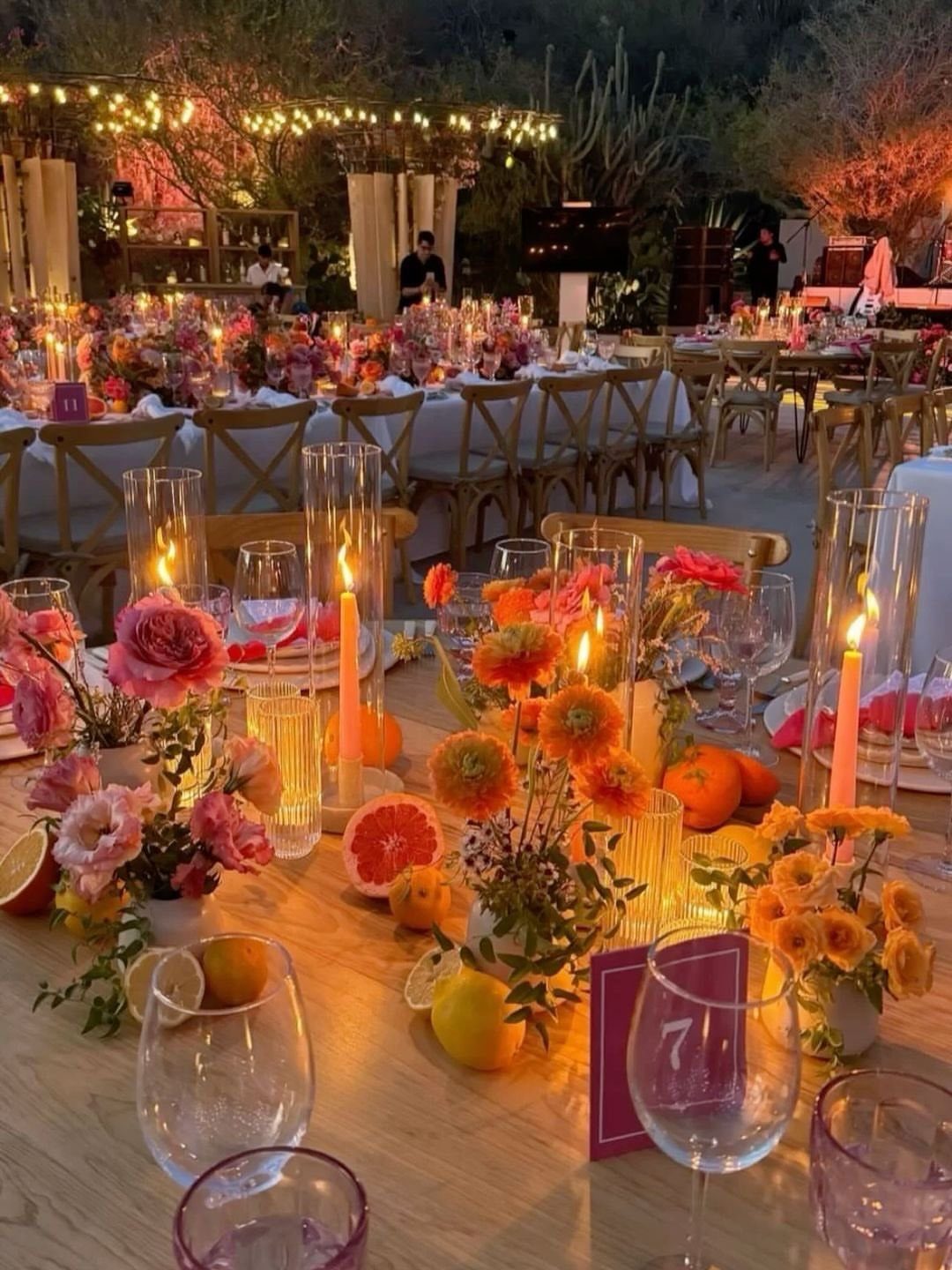 Wedding table setting with colorful flowers, citrus fruits, and glowing candles under evening lights.