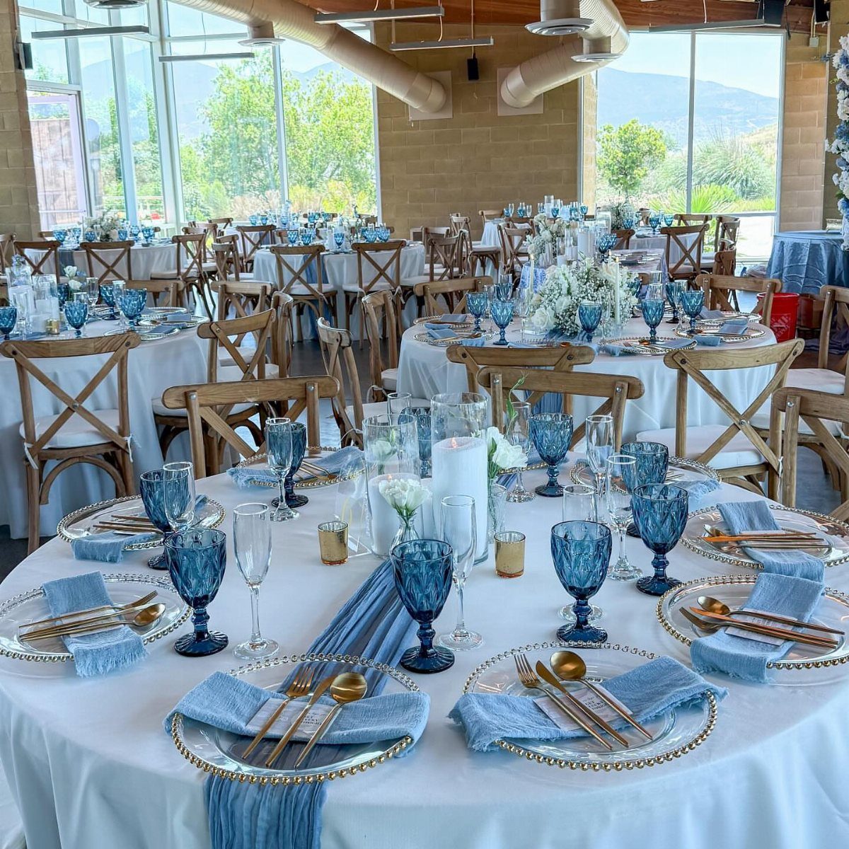 Wedding table setting featuring dusty blue napkins, glassware, and white floral centerpieces on white linens.