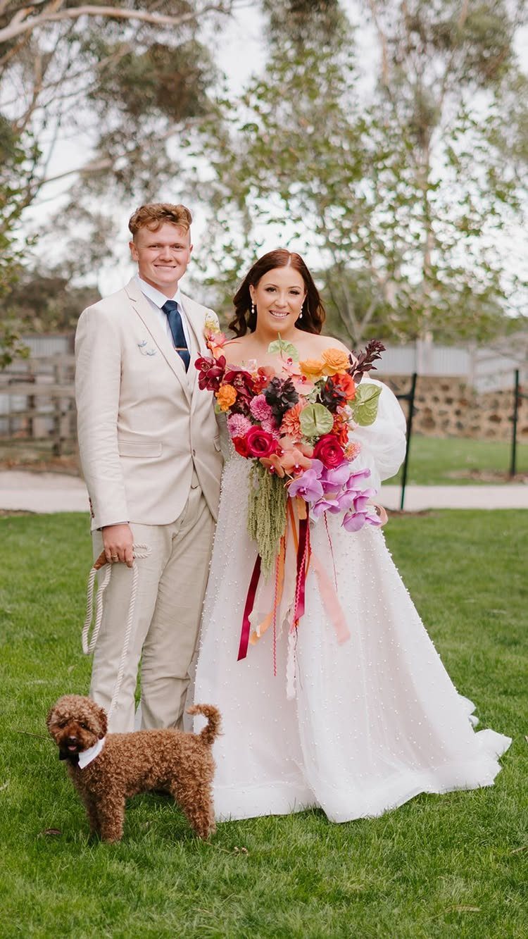 A bride holding a colorful floral bouquet stands next to a groom and a small brown dog.