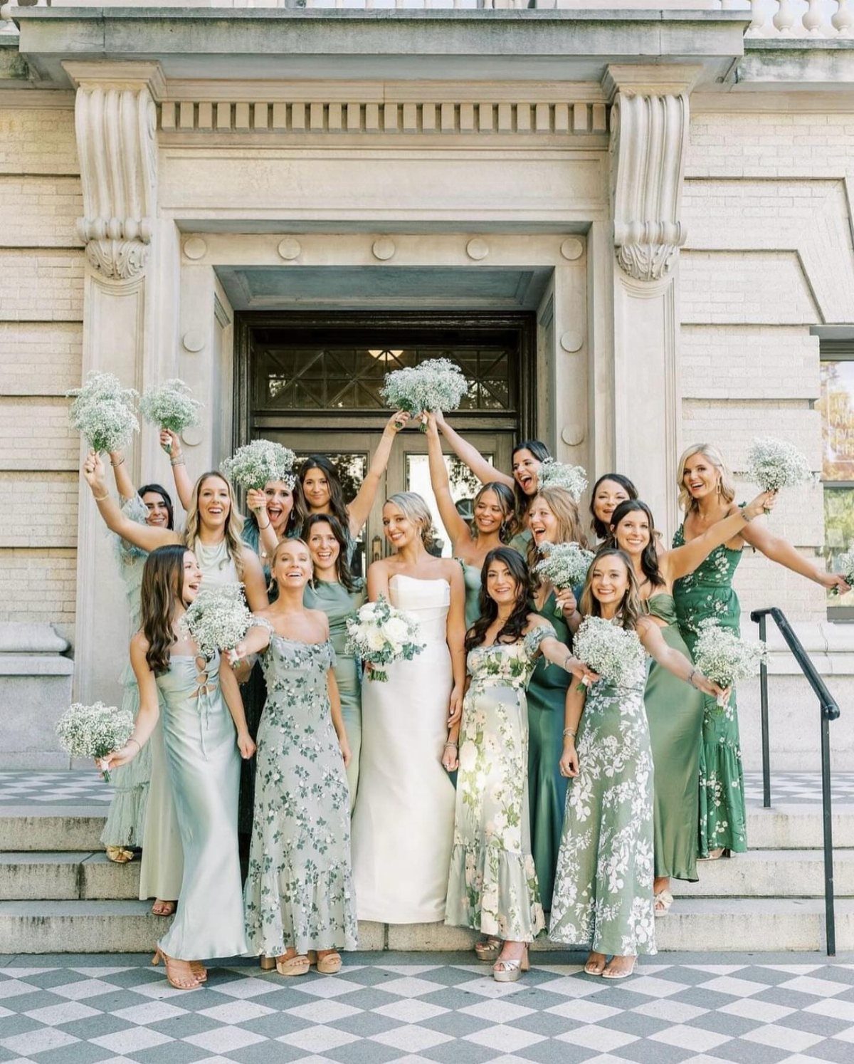 Bridesmaids in sage green dresses holding white bouquets standing on stone steps with a bride.