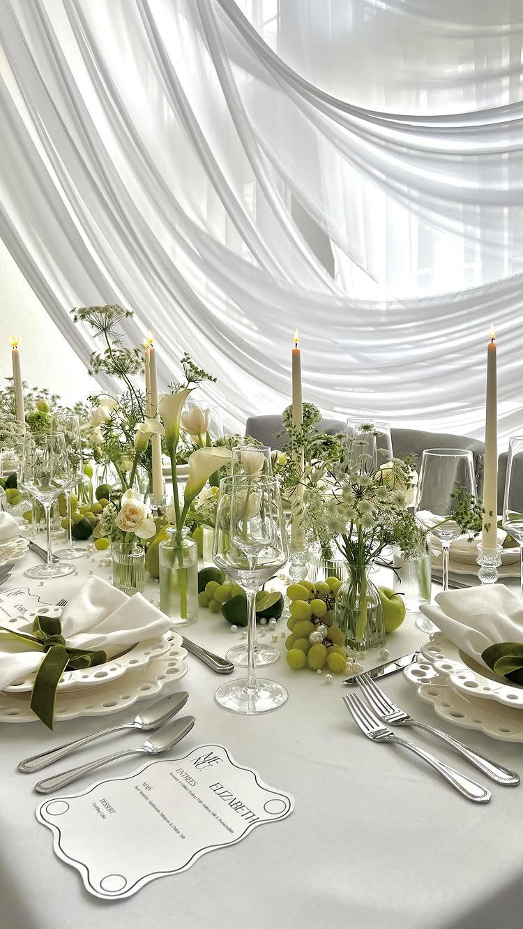 A wedding table decorated with white flowers, green foliage, grapes, and lit taper candles.
