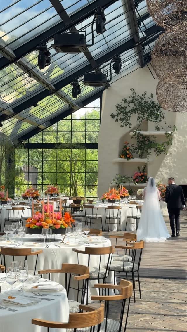 Wedding reception tables with bright orange floral centerpieces in a bright glass-walled venue.