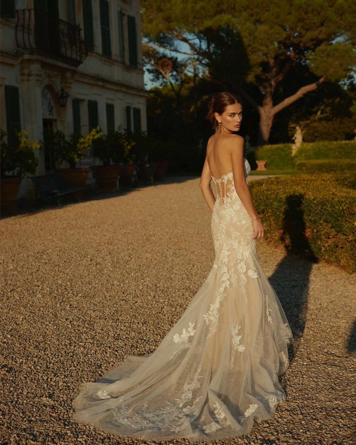 Tight Lace Wedding Dress: 8 Fitted Styles to Shape the Day 4 A woman wearing a white lace mermaid wedding dress with an open back stands on a gravel path.