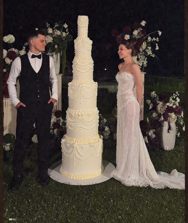 Tight Lace Wedding Dress: 8 Fitted Styles to Shape the Day 7 A bride in a lace wedding dress stands beside a groom and a tall tiered cake.