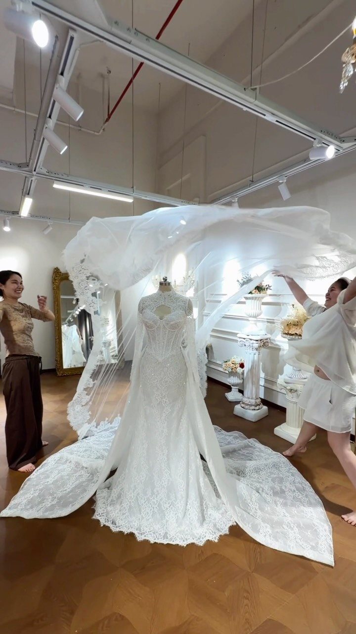 Tight Lace Wedding Dress: 8 Fitted Styles to Shape the Day 1 A white lace wedding dress on a mannequin with a flowing sheer veil being held by two women.