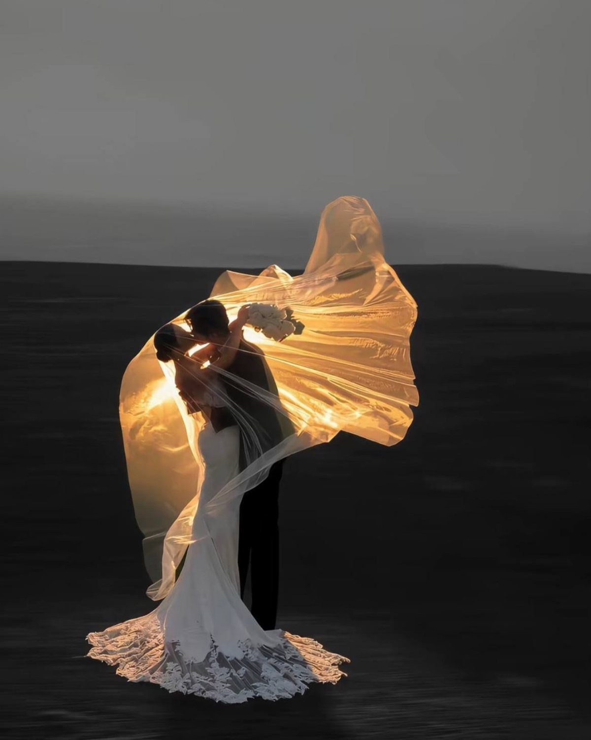 A bride and groom embrace under a glowing, backlit veil at dusk.